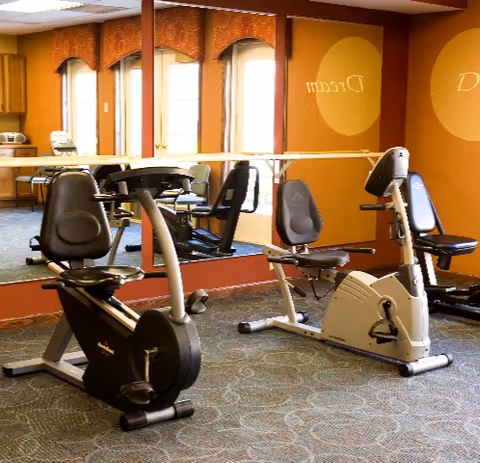 A small fitness room with two stationary exercise bikes positioned on a carpeted floor. The room has large mirrors on one wall reflecting the equipment and windows with orange valances. The walls are painted orange with circular designs and the word 'Dream' partially visible.