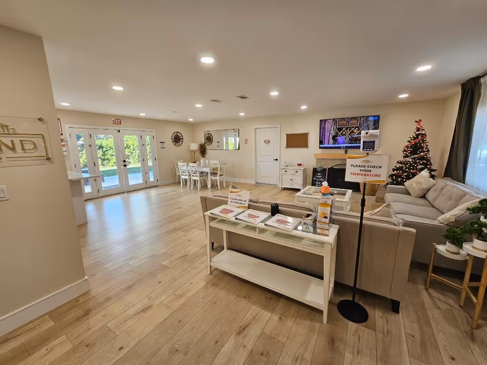 A spacious, well-lit common area in a senior living facility with light wood flooring and beige walls. The room features a sitting area with beige sofas, a coffee table, and a decorated Christmas tree. A television is mounted above a fireplace. There is a dining area with a table and chairs near glass double doors leading outside. A sign on a stand asks visitors to check their temperature. Various informational signs and hand sanitizer are placed on a console table behind the sofas.