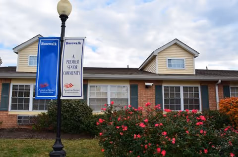 Exterior view of a single-story brick building with beige siding and multiple windows. In front of the building, there is a garden with blooming pink flowers and green shrubs. A black lamppost stands near the garden with two vertical signs that read 'Rosewalk' and 'A Premier Senior Community' along with the American Senior Communities logo. The sky is partly cloudy.