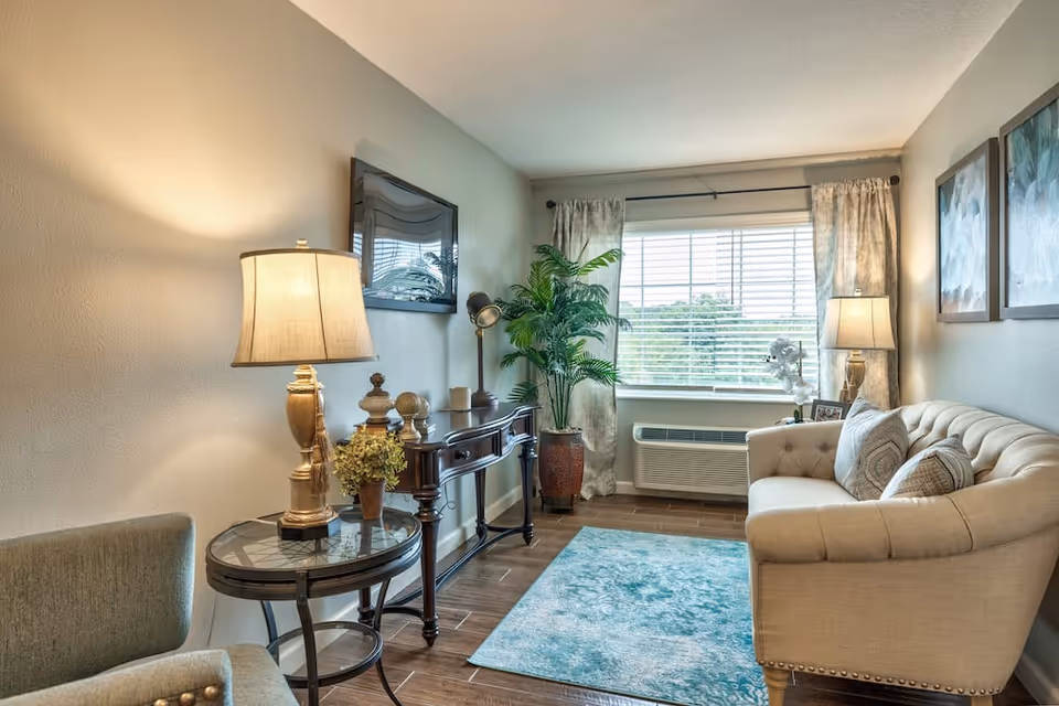 A cozy living room with a beige tufted sofa adorned with patterned pillows, a blue area rug on wooden flooring, a round glass-top side table with a lamp and small plant, a dark wooden console table with decorative items, a tall potted plant near a window with blinds and curtains, and two framed artworks on the wall.