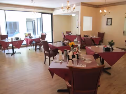 A dining room with several tables covered in burgundy tablecloths, each set with plates, cups, and condiments. The room has wooden flooring, beige walls, chandeliers, and large windows letting in natural light.