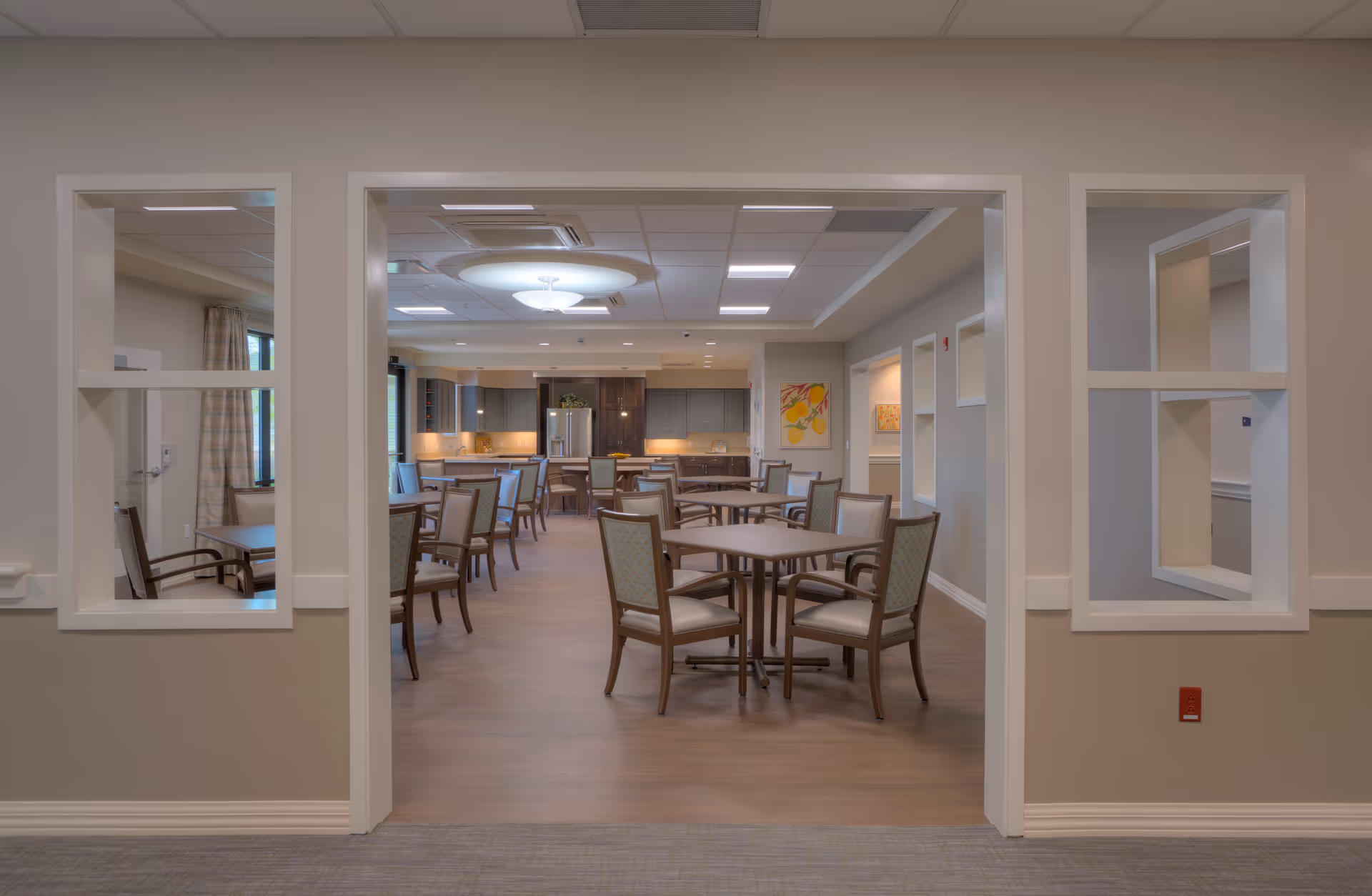 Open dining room with multiple tables and chairs facing a kitchen area in a senior living facility.