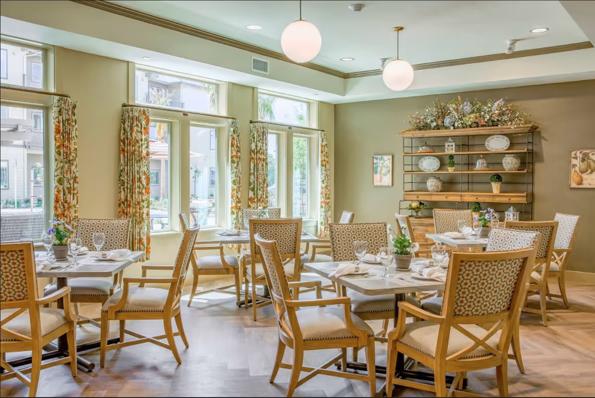 A bright dining room with several wooden tables and cushioned chairs arranged neatly. The room features large windows with floral curtains allowing natural light to fill the space. There are decorative plants on the tables and a wooden shelving unit on the wall adorned with plates, small plants, and other decorative items. Two round pendant lights hang from the ceiling.
