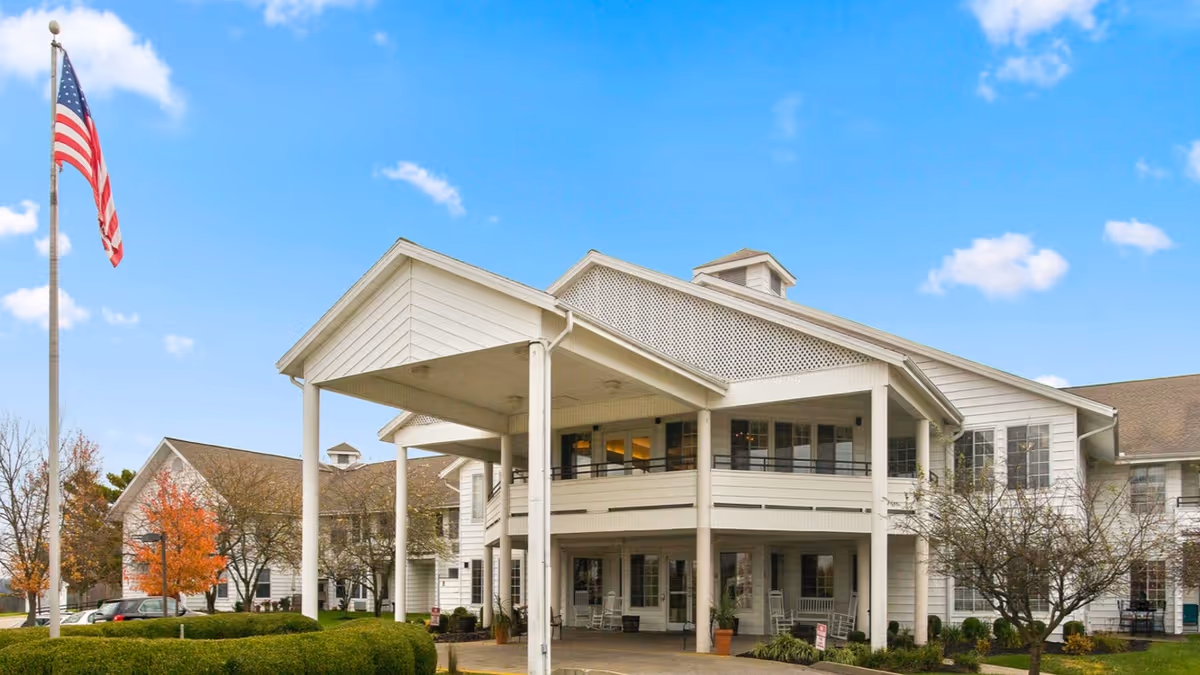 Exterior view of a senior living facility with white siding and a covered entrance supported by columns. There is an American flag on a flagpole to the left, some trees with autumn foliage, and a clear blue sky with a few clouds.