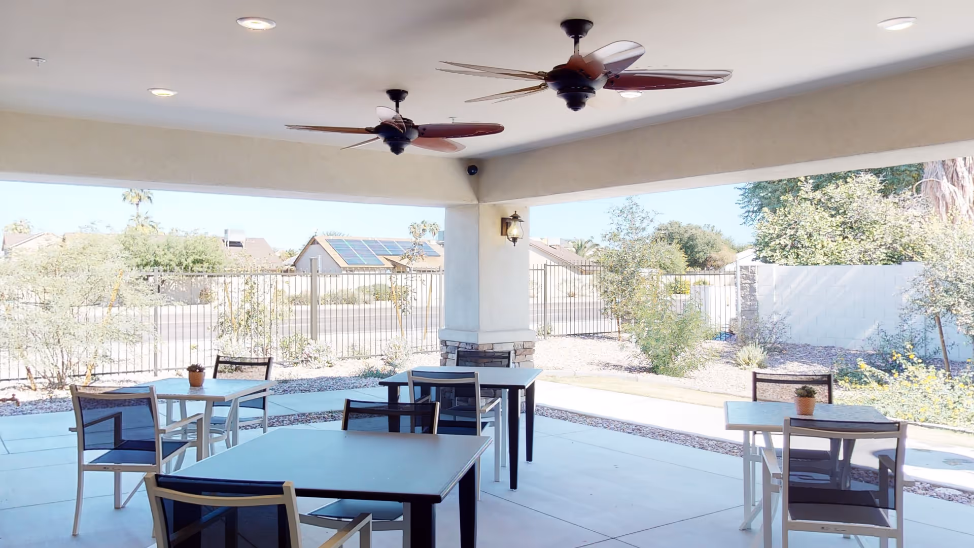 Covered outdoor patio area with several tables and chairs arranged for seating. The patio has ceiling fans and overlooks a fenced garden area with shrubs and trees, with houses visible in the background under a clear sky.