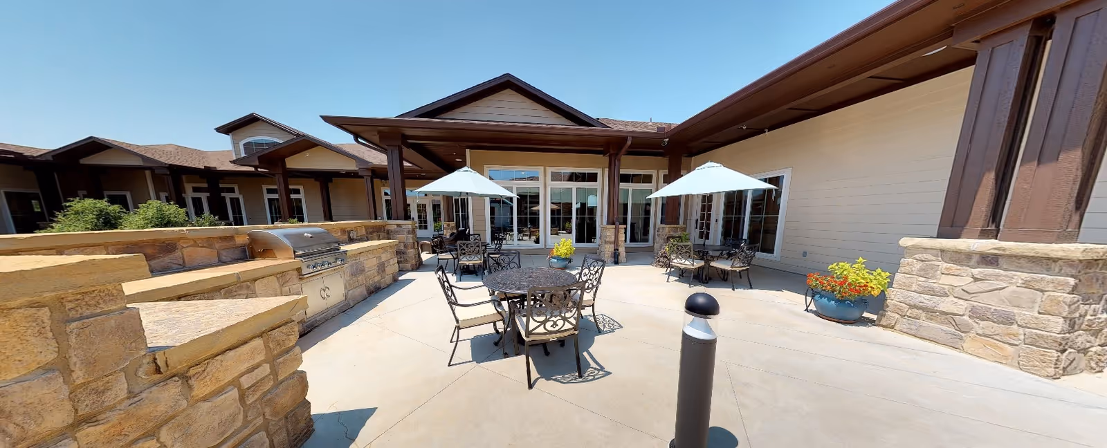 Outdoor patio area at Grand Brook Memory Care of Zionsville featuring multiple round metal tables with chairs and umbrellas, stone walls, a built-in stainless steel grill, potted plants, and a covered porch with large windows and doors leading inside.