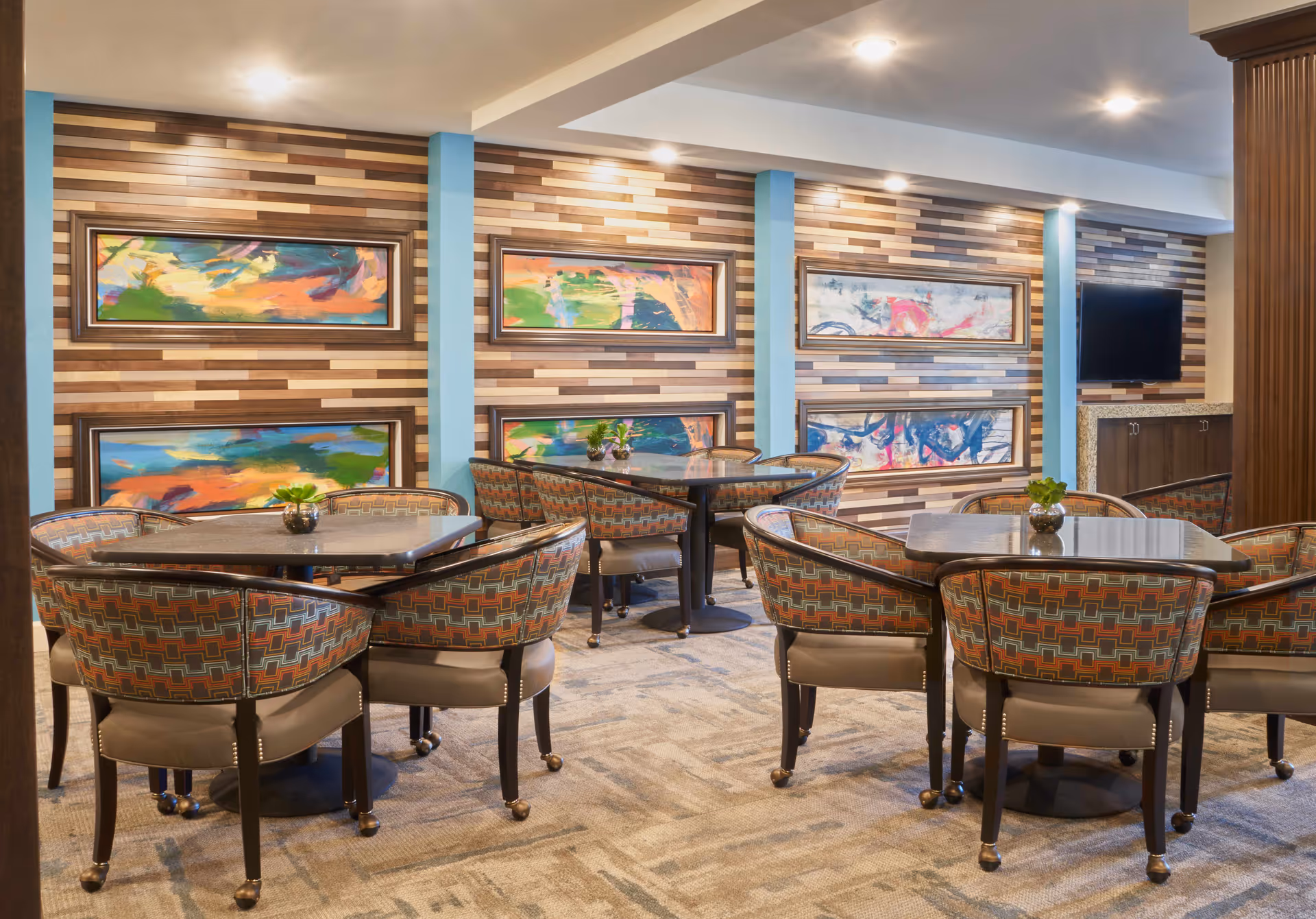 A cozy dining area with several square tables surrounded by cushioned chairs featuring a geometric pattern. The walls are decorated with horizontal wooden panels and colorful abstract paintings. There is a flat-screen TV mounted on the wall to the right.