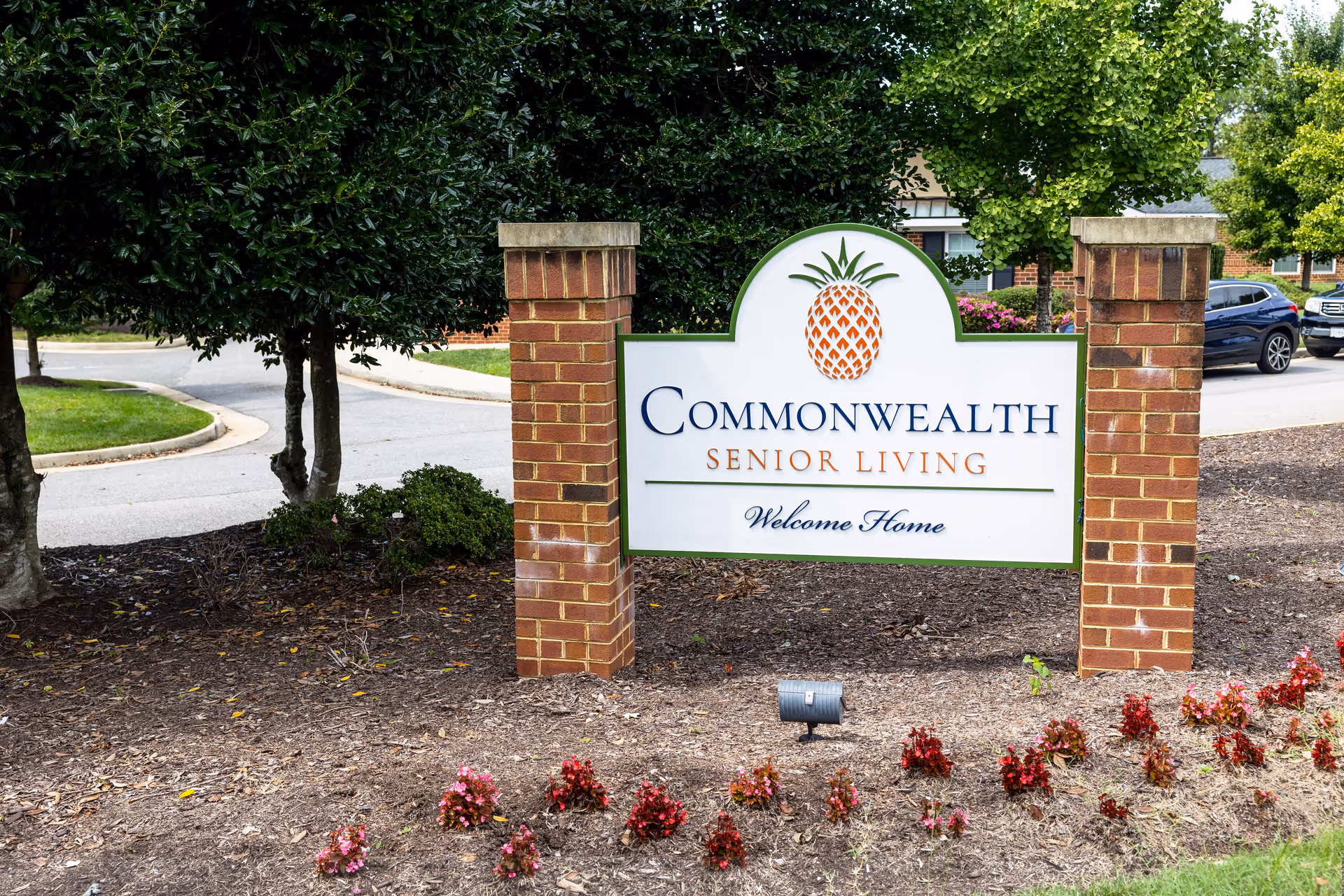 A brick and white sign with a pineapple logo that reads 'Commonwealth Senior Living Welcome Home' situated outdoors with trees, a curved road, and parked cars in the background.