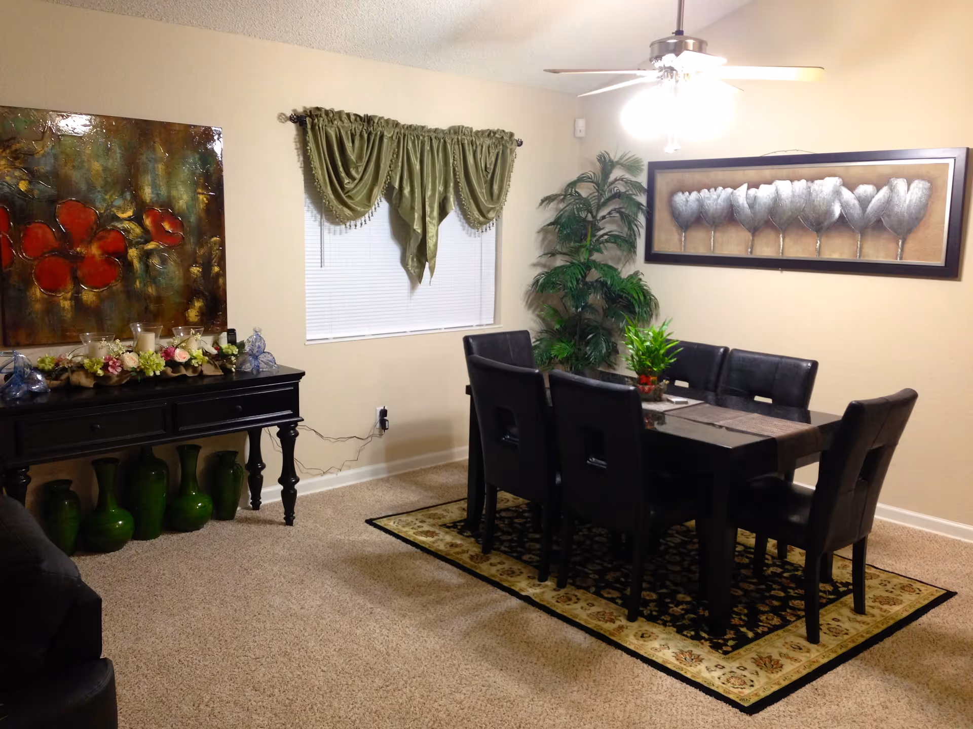 A dining room with a rectangular dark wooden table surrounded by six black leather chairs. The table is set on a decorative area rug with floral patterns. On the table, there is a small green plant centerpiece. The room has beige walls with two large framed artworks: one with red flowers on the left wall and another with silver tulip-like flowers on the right wall. A window with green drapes is centered on the back wall, and a tall green potted plant is placed in the corner. A ceiling fan with lights hangs above the table. A black console table with decorative items and green vases underneath is positioned against the left wall.