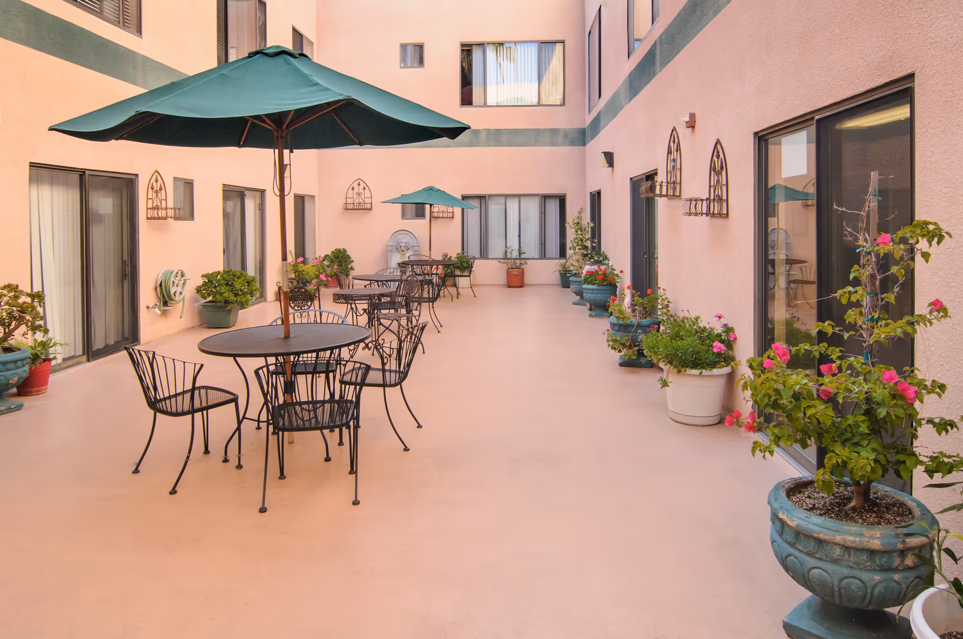Outdoor courtyard area with several black metal tables and chairs, each table shaded by a green umbrella. The courtyard is surrounded by a pink building with multiple windows and sliding glass doors. Various potted plants and flowers line the edges of the courtyard.