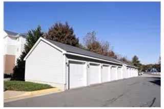 A row of white garage doors attached to a single building with a sloped roof, situated next to a paved driveway. Trees and part of a multi-story building are visible in the background under a clear blue sky.