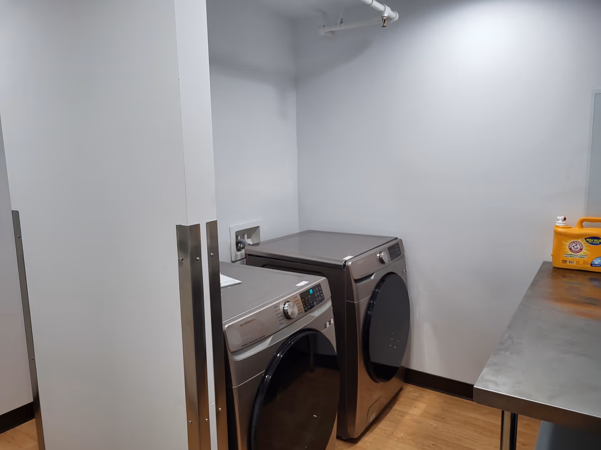 Laundry room with a front-loading washing machine and dryer side by side, a stainless steel countertop on the right with a yellow container of Arm & Hammer laundry detergent, and white walls with a wooden floor.