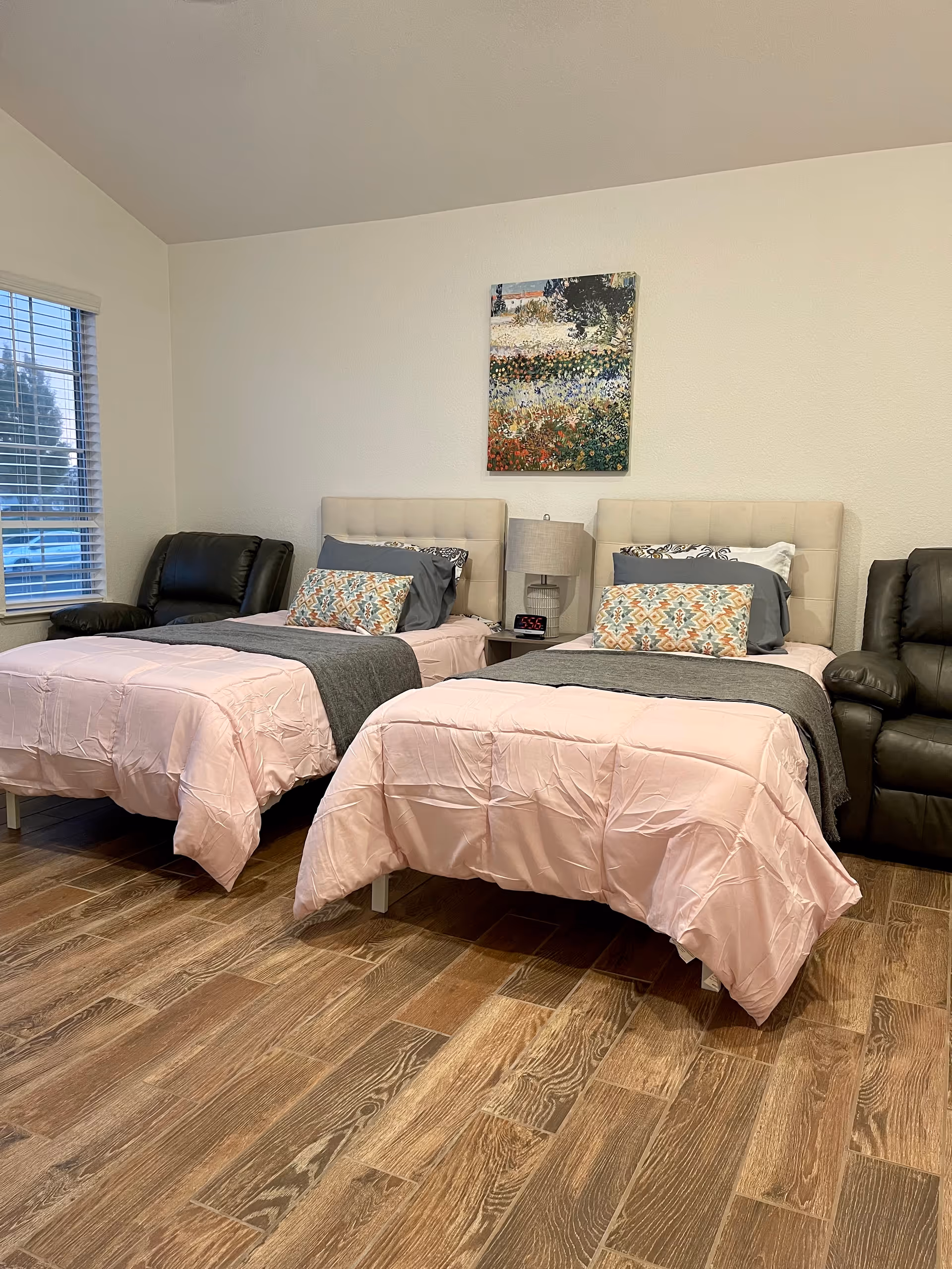 A bedroom with two twin beds side by side, each with light pink comforters, gray blankets, and decorative pillows. Between the beds is a nightstand with a lamp and a digital alarm clock. There are two black recliner chairs on either side of the beds. A window with blinds is on the left wall, and a colorful painting hangs above the nightstand on a plain white wall. The floor is wooden.