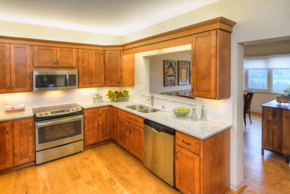 A bright kitchen with wooden cabinets, stainless steel appliances including a microwave, oven, and dishwasher. The countertops are light-colored with a bowl of green apples and decorative items. There is a pass-through window opening to an adjacent room with artwork on the wall and a dining area visible through a doorway.