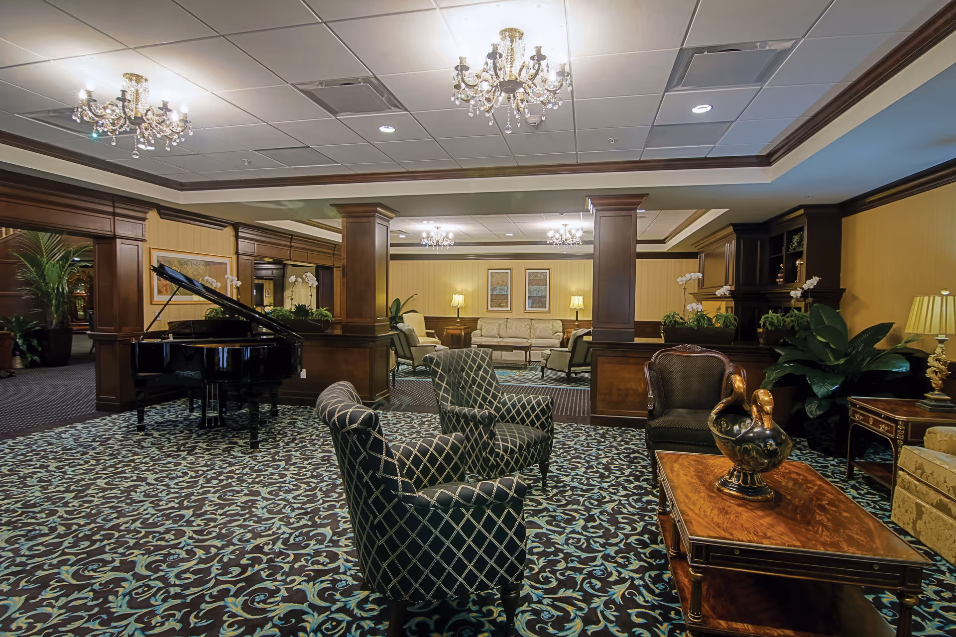 A spacious and elegantly decorated living room area with patterned carpet, two chandeliers, dark wood paneling, and several seating arrangements including armchairs, sofas, and a grand piano. There are plants and framed artwork on the walls, creating a warm and inviting atmosphere.