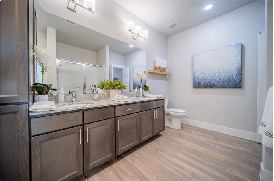 A modern bathroom with a long granite countertop featuring two sinks, wooden cabinets underneath, and decorative plants. There is a large mirror above the sinks with wall-mounted light fixtures. A toilet is visible in the corner next to a wall-mounted shelf holding towels. The bathroom has wood-look flooring and a framed abstract painting on the wall.