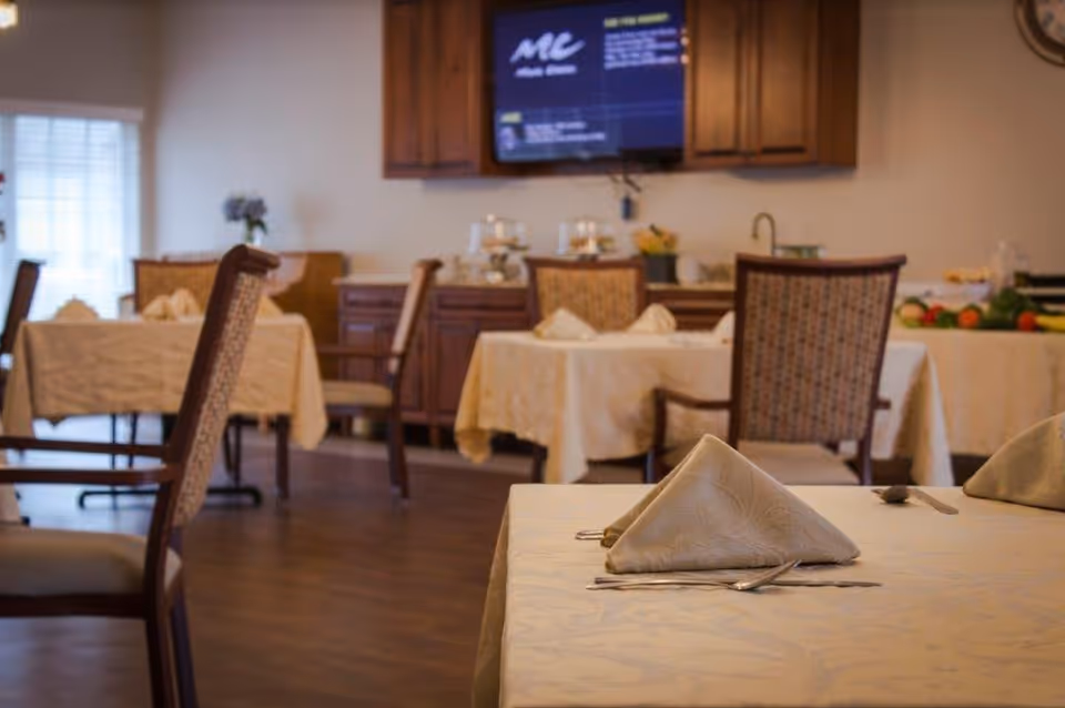 Dining room with tables covered in white tablecloths, each set with folded napkins and silverware. Wooden chairs with patterned upholstery surround the tables. In the background, there is a wooden cabinet with a television mounted above it and a buffet table with food items.