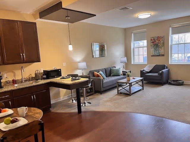 A cozy living area in a senior living facility featuring a gray sofa with colorful cushions, a matching armchair, a coffee table with a plant, and two windows letting in natural light. The room has beige walls and carpeted flooring. Adjacent to the living area is a small kitchen with dark wood cabinets, a granite countertop, a microwave, and a small dining table with chairs.