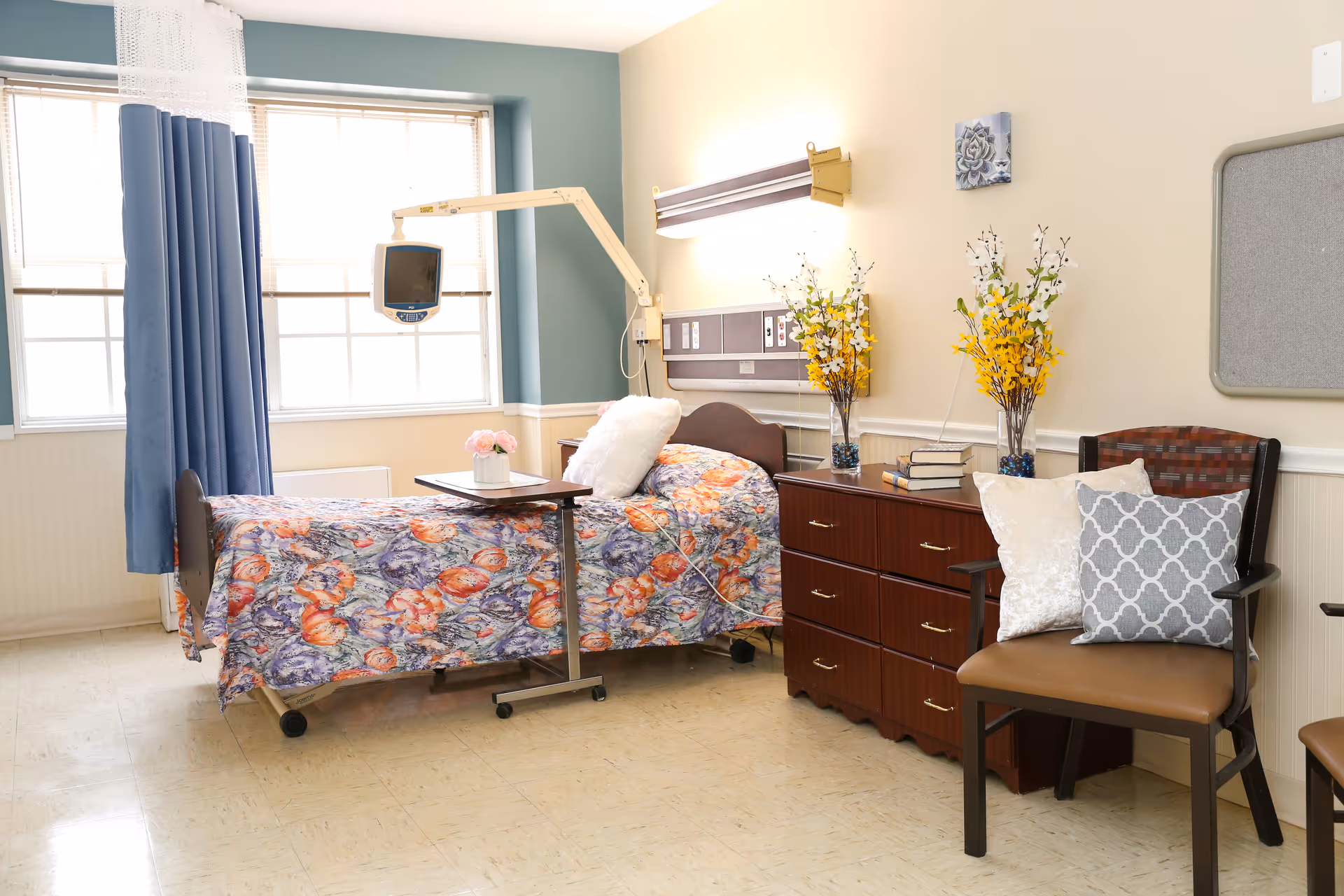 A well-lit room with a hospital bed covered in a colorful floral blanket, a small overbed table with a flower vase, a wooden dresser with books and flower arrangements, and a chair with decorative pillows. The room has large windows with blue curtains and medical equipment mounted on the wall.