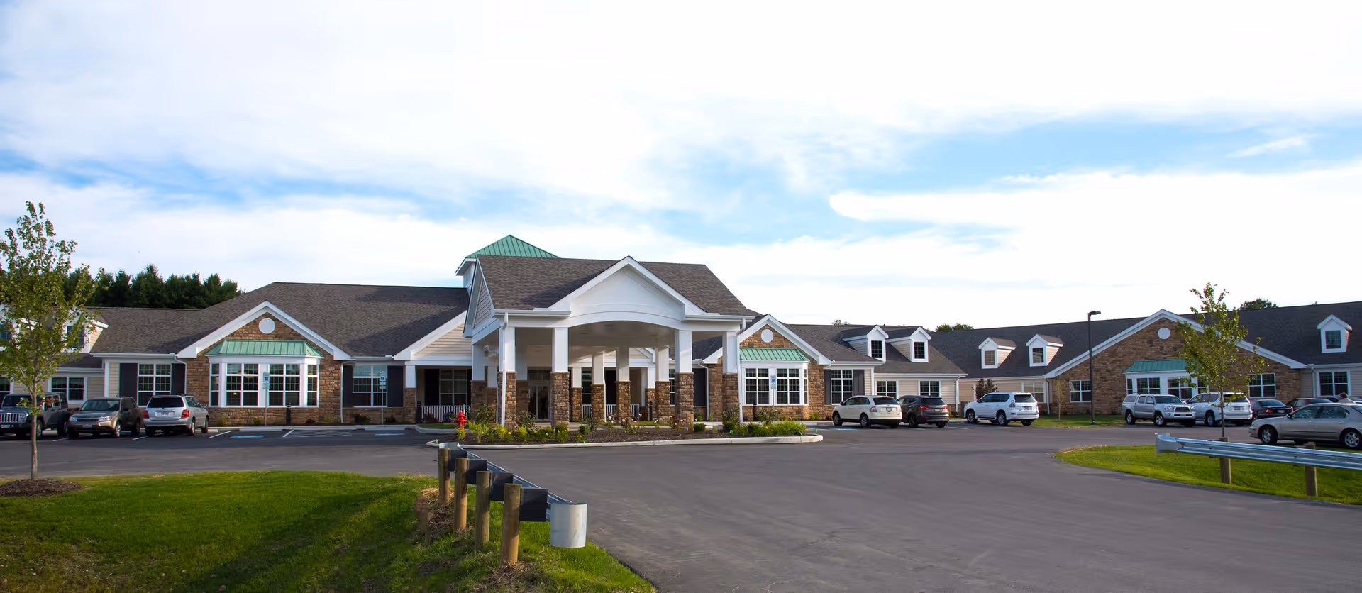 Front entrance of a single-story senior living facility with a porte-cochère, parked cars, and landscaped grounds.