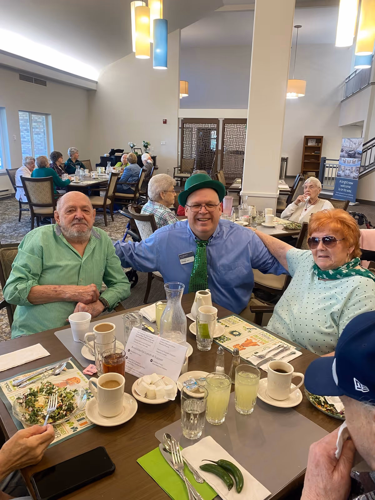 A group of elderly people sitting around a dining table in a senior living facility, enjoying a meal together. One man in the center is wearing a green hat and green tie, smiling with his arms around two other seniors. The table has cups, glasses with drinks, plates with food, and condiments. Other seniors are visible in the background seated at tables in a well-lit dining room with high ceilings and modern light fixtures.