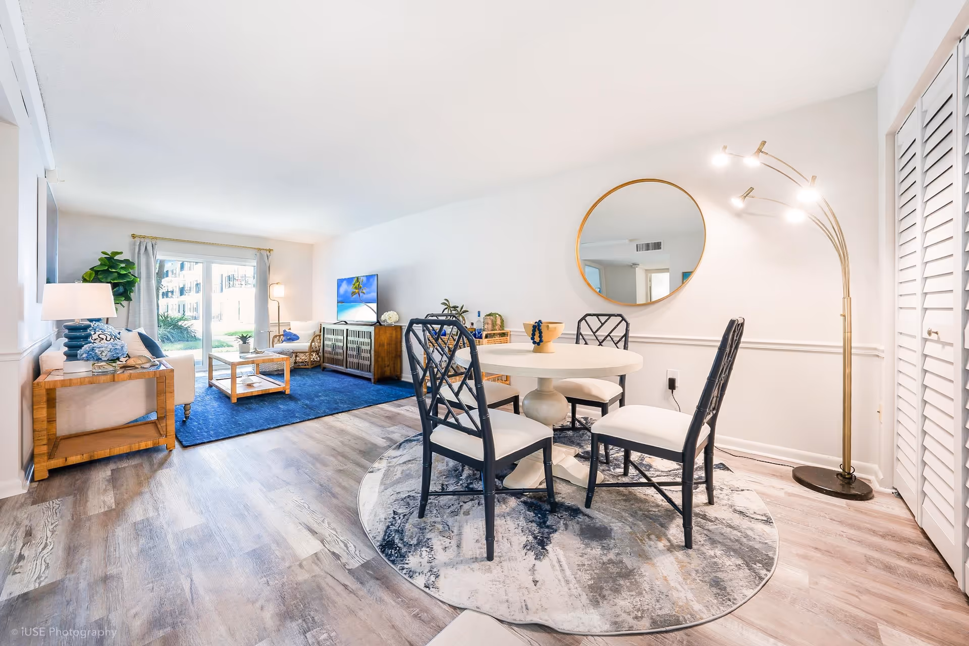 Bright and modern senior living interior featuring a dining area with a round table and four chairs on a patterned rug, a large round mirror on the wall, and a stylish floor lamp. The living area includes a blue rug, a wooden coffee table, a TV on a wooden console, a sofa, and a large window with curtains letting in natural light.
