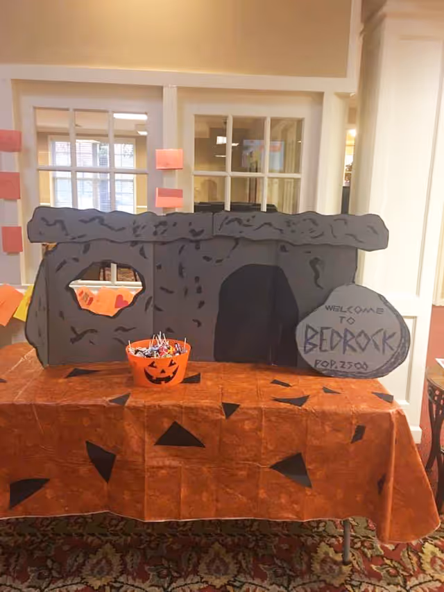 A Halloween-themed display resembling the Flintstones' Bedrock house made from cardboard and placed on a table covered with an orange cloth decorated with black shapes. A small orange pumpkin bucket filled with candy is in front of the display. The background shows windows and interior walls of a senior living facility.