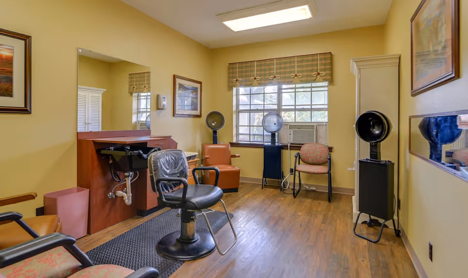A small salon room with a black salon chair in front of a mirror and sink, two hair dryer chairs near a window with an air conditioner, and framed artwork on the yellow walls.