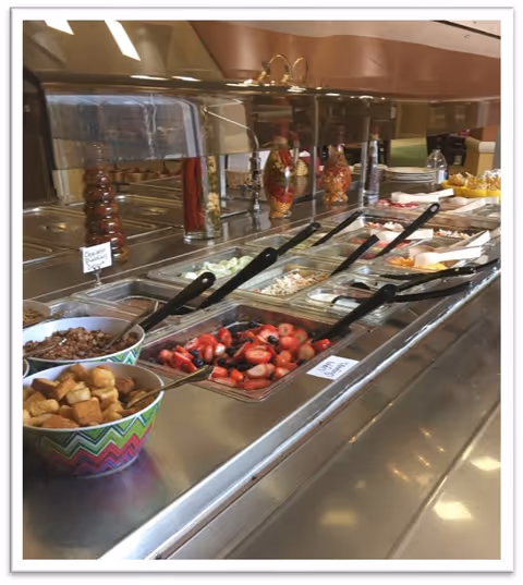 Buffet-style salad bar with various fresh ingredients including strawberries, croutons, chopped vegetables, and salad dressings in a retirement community dining area.