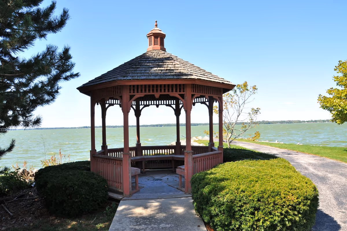 A wooden gazebo with a shingled roof situated near a body of water, surrounded by green bushes and trees under a clear blue sky.