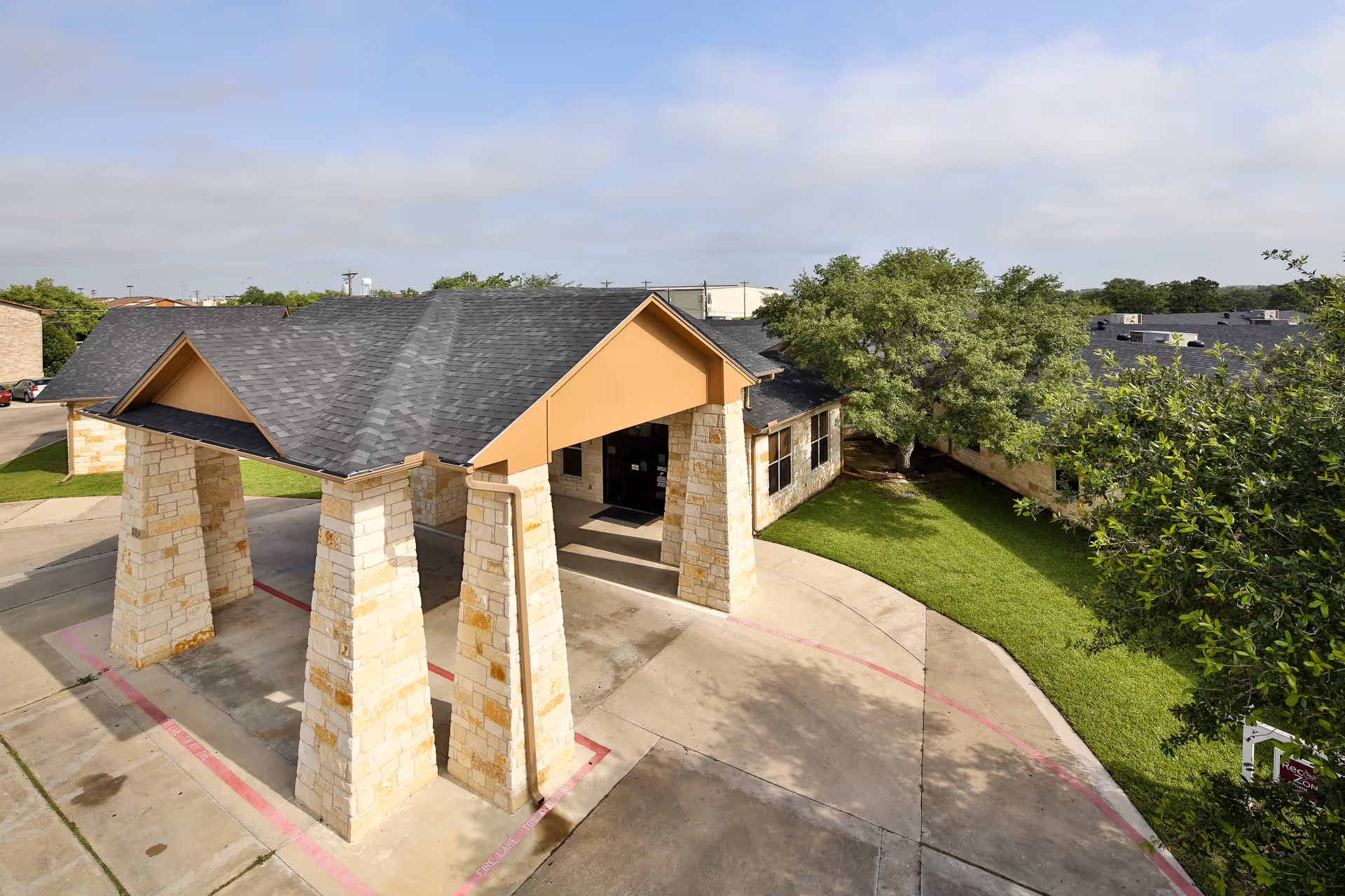Covered entrance with stone columns and driveway leading to a single-story building surrounded by grass and trees.