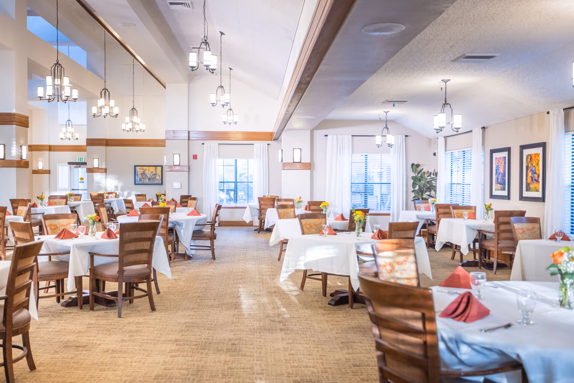 A bright and spacious dining room with multiple tables covered in white tablecloths, each set with folded red napkins, glassware, and small flower arrangements. The room features wooden chairs, large windows with white curtains, warm lighting from hanging chandeliers, and framed artwork on the walls.