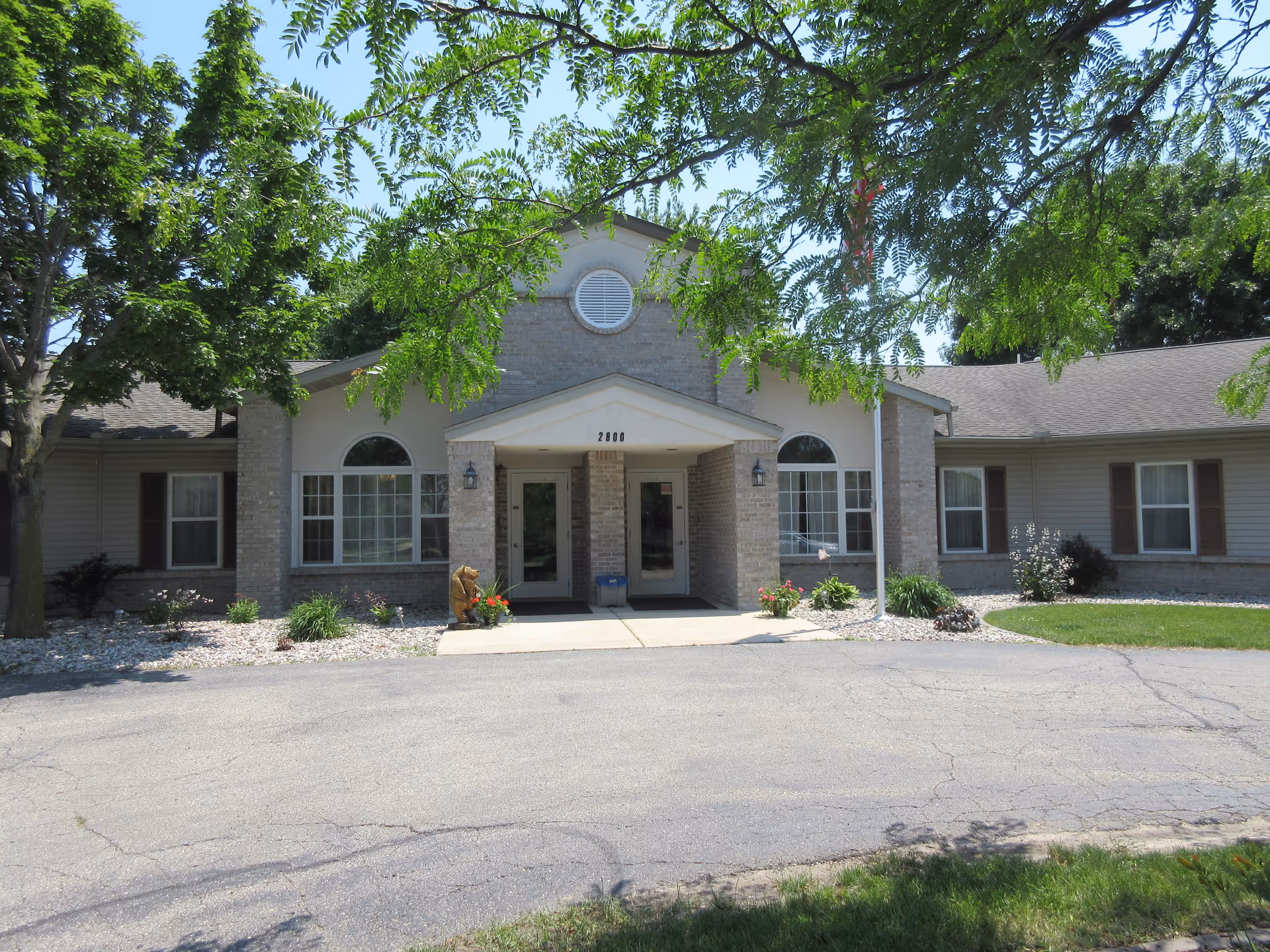 Front exterior view of a single-story building with beige brick and siding, featuring two glass entrance doors under a small peaked roof. The building is surrounded by landscaped areas with small plants and trees, and a paved driveway in front. The address number 2800 is visible above the entrance.