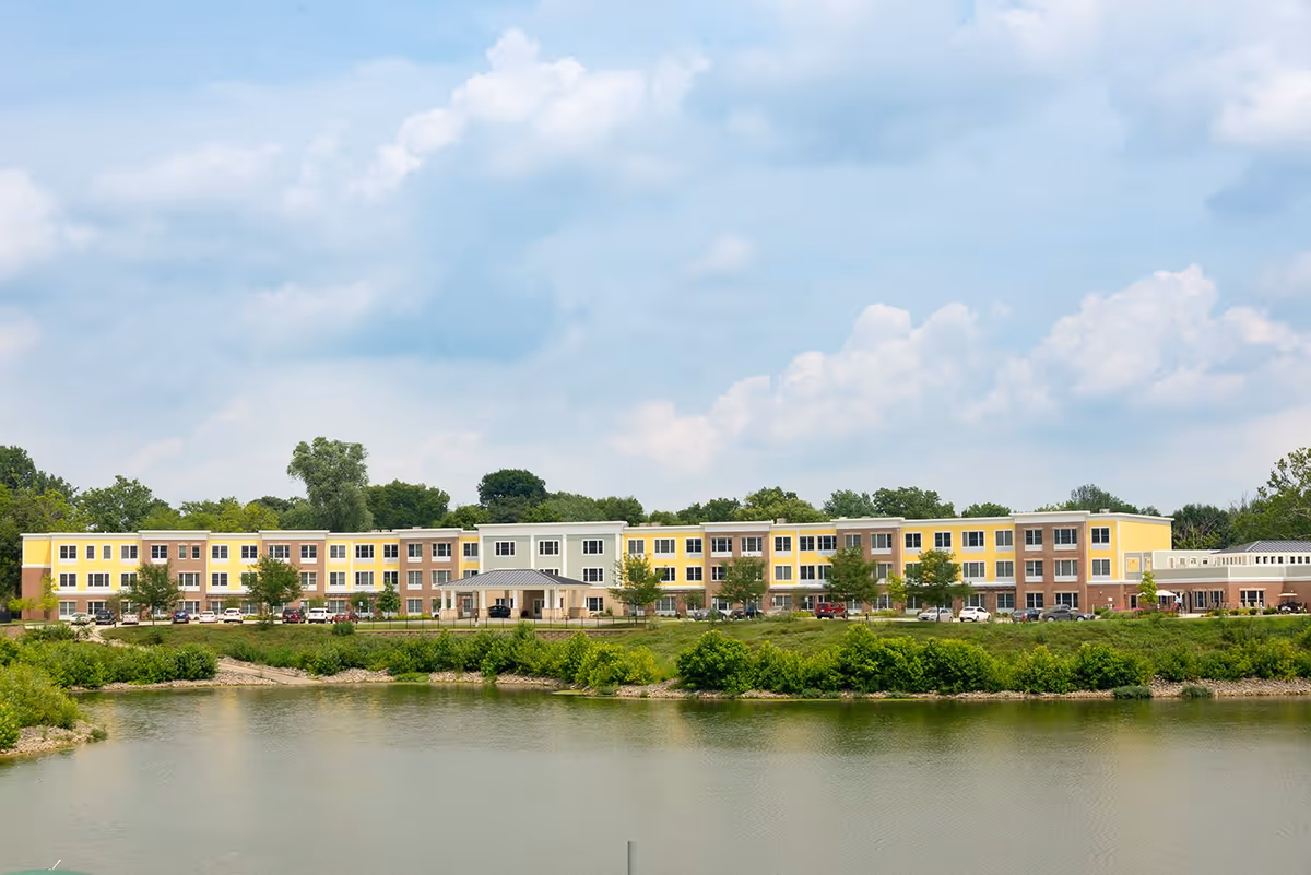 Wide exterior view of Traditions at Solana, a three-story senior living facility with a mix of yellow, beige, and brick facade, situated behind a pond with greenery and trees under a partly cloudy sky.