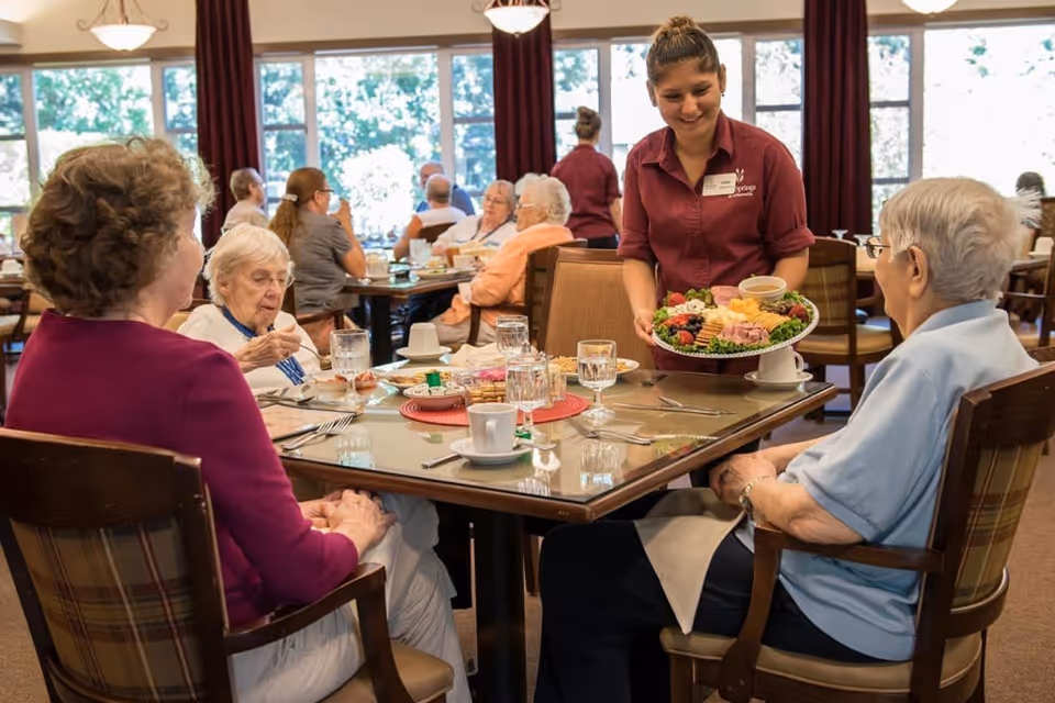 A dining room in a senior living facility where elderly residents are seated around tables enjoying a meal. A staff member wearing a maroon uniform is serving a platter of assorted food including fruits, cheese, and meats to one of the residents. The room has large windows with natural light and red curtains.