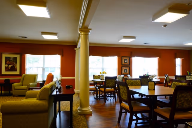 Interior view of a senior living facility common area with multiple tables and chairs arranged for dining or socializing. The room features warm orange walls, large windows letting in natural light, framed artwork on the walls, and comfortable armchairs near a column dividing the space.