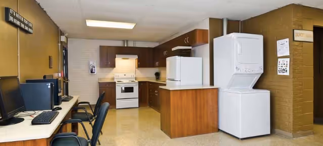 Interior view of a multipurpose room featuring a kitchen area with wooden cabinets, a white stove, refrigerator, and a stacked washer and dryer. On the left side, there is a row of desks with computers and chairs. The walls are painted brown and beige, and there is a sign on the wall above the computers.
