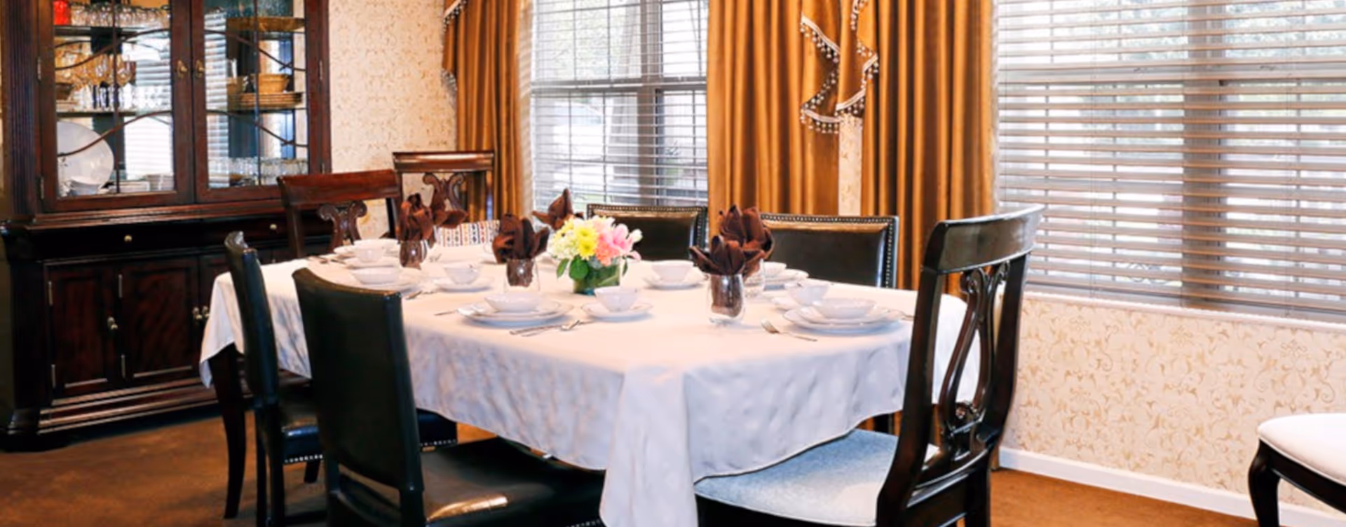 A dining room with a rectangular table covered with a white tablecloth, set with white dishes, bowls, cups, and silverware. Brown napkins are folded and placed in glasses on the table. A small flower arrangement is in the center. The room has large windows with blinds and gold curtains, and a dark wooden china cabinet filled with dishes and glassware is against the wall.