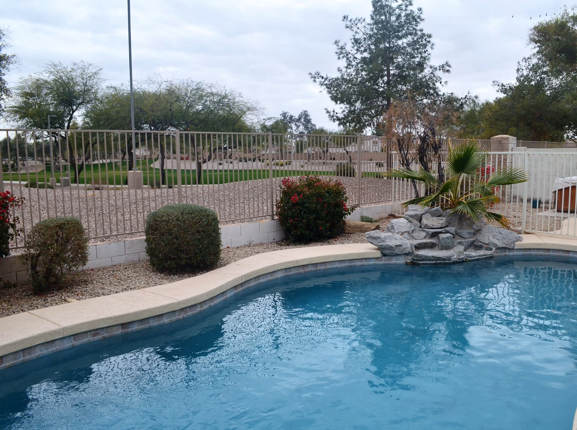 Outdoor swimming pool with clear blue water, surrounded by a beige concrete edge. There are small bushes and a rock waterfall feature with a palm plant near the pool. A metal fence encloses the area, and beyond the fence, there are trees and a grassy area under a cloudy sky.
