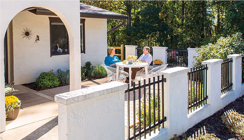 Two people sitting at a round white table on a patio outside a white stucco building with a black metal fence and greenery surrounding the area.