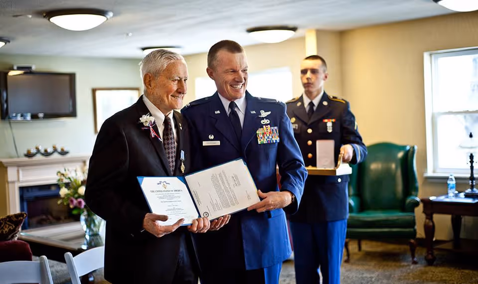 An elderly man in a suit with a flower boutonniere is smiling and holding a certificate alongside a man in a decorated military uniform. Another man in military uniform stands in the background holding a box. The setting is a well-lit room with chairs, a green armchair, a table with a water bottle, and a fireplace with flowers.