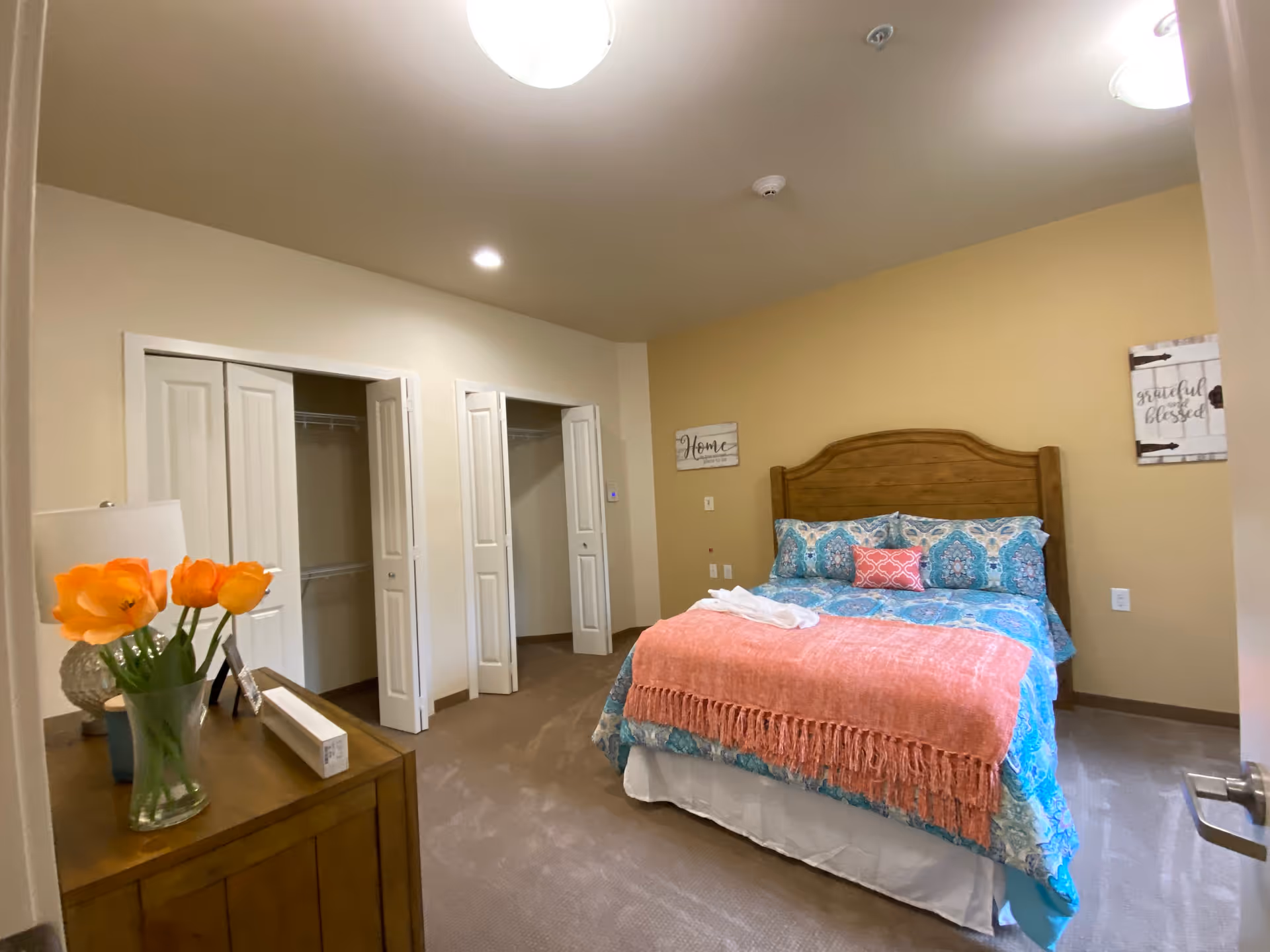 A bedroom with a wooden headboard bed dressed in blue and pink patterned bedding and a coral throw blanket. There are two open closets with white bi-fold doors on the left wall. A wooden dresser with a vase of orange flowers and a lamp is visible in the foreground. The walls are painted beige with two decorative signs hanging on the wall.