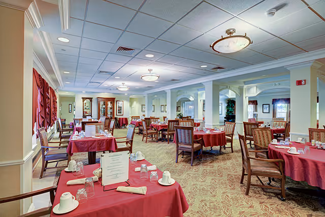 A spacious dining room in a senior living facility with multiple round tables covered in red tablecloths, each set with white cups, glasses, and napkins. The room features beige walls, carpeted floors, decorative columns, and ceiling lights. Large windows with red curtains allow natural light to enter the room.