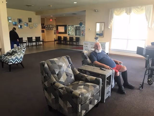 A spacious common area in an assisted living facility with patterned armchairs and a small table. A man with a beard, wearing a blue shirt and red shorts, is seated in one of the chairs. Another person is seen walking in the background near a wall with framed pictures. Large windows with sheer curtains allow natural light to fill the room.
