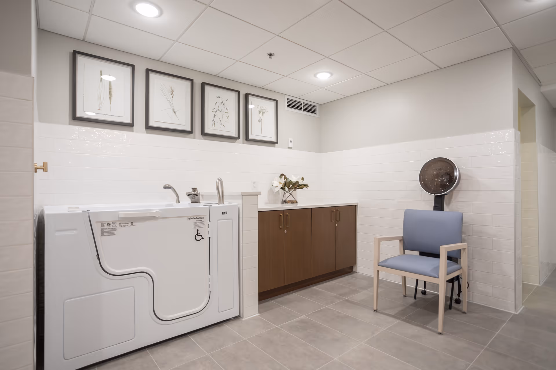 Accessible walk-in bathtub and sink next to wooden cabinets and a chair with a hooded hair dryer in a tiled room with framed botanical prints.
