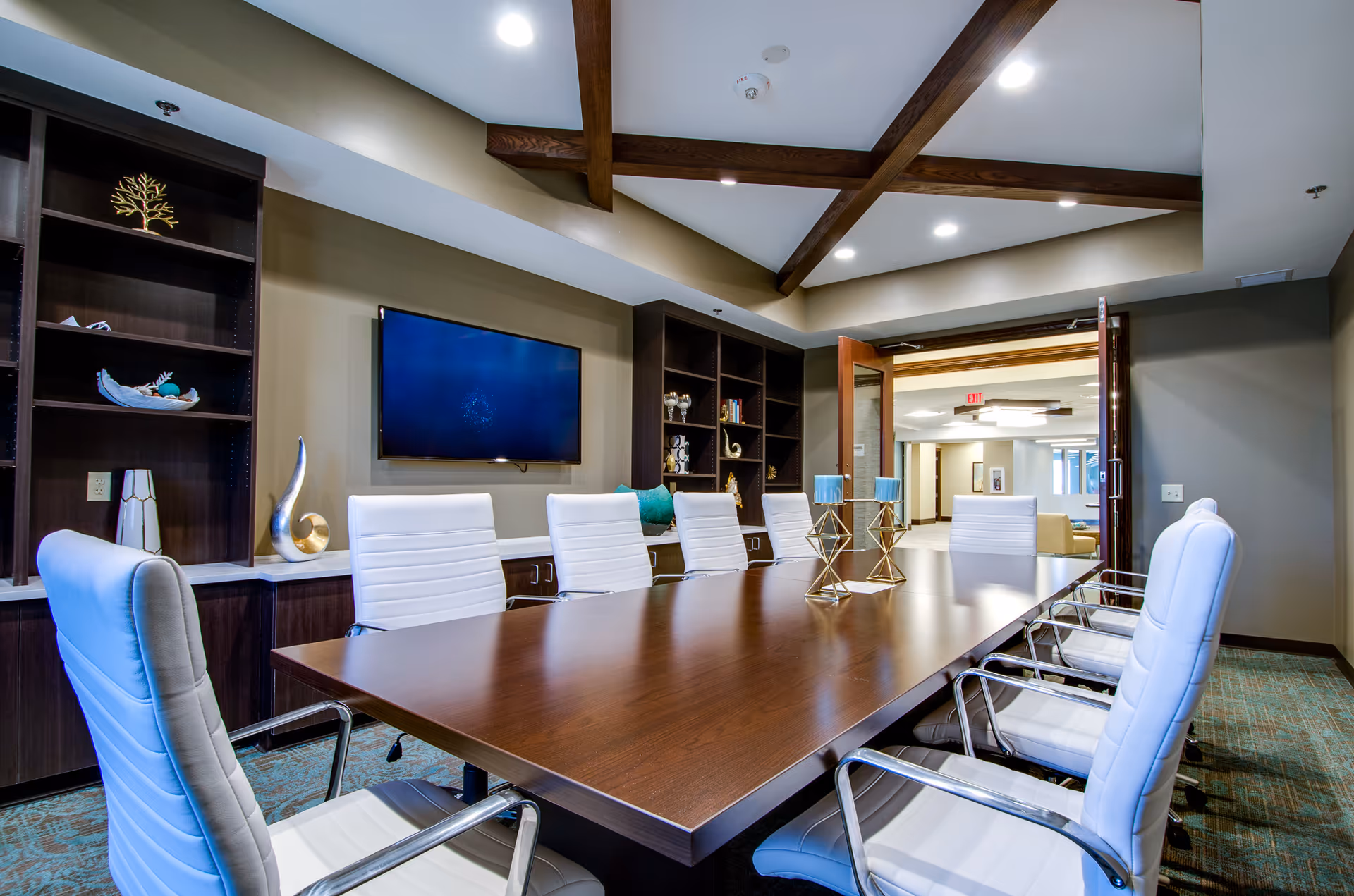 A modern conference room with a large wooden table surrounded by white leather chairs. The room features dark wood built-in shelves with decorative items, a flat-screen TV mounted on the wall, and exposed wooden beams on the ceiling. The door is open, showing a hallway and another seating area outside.