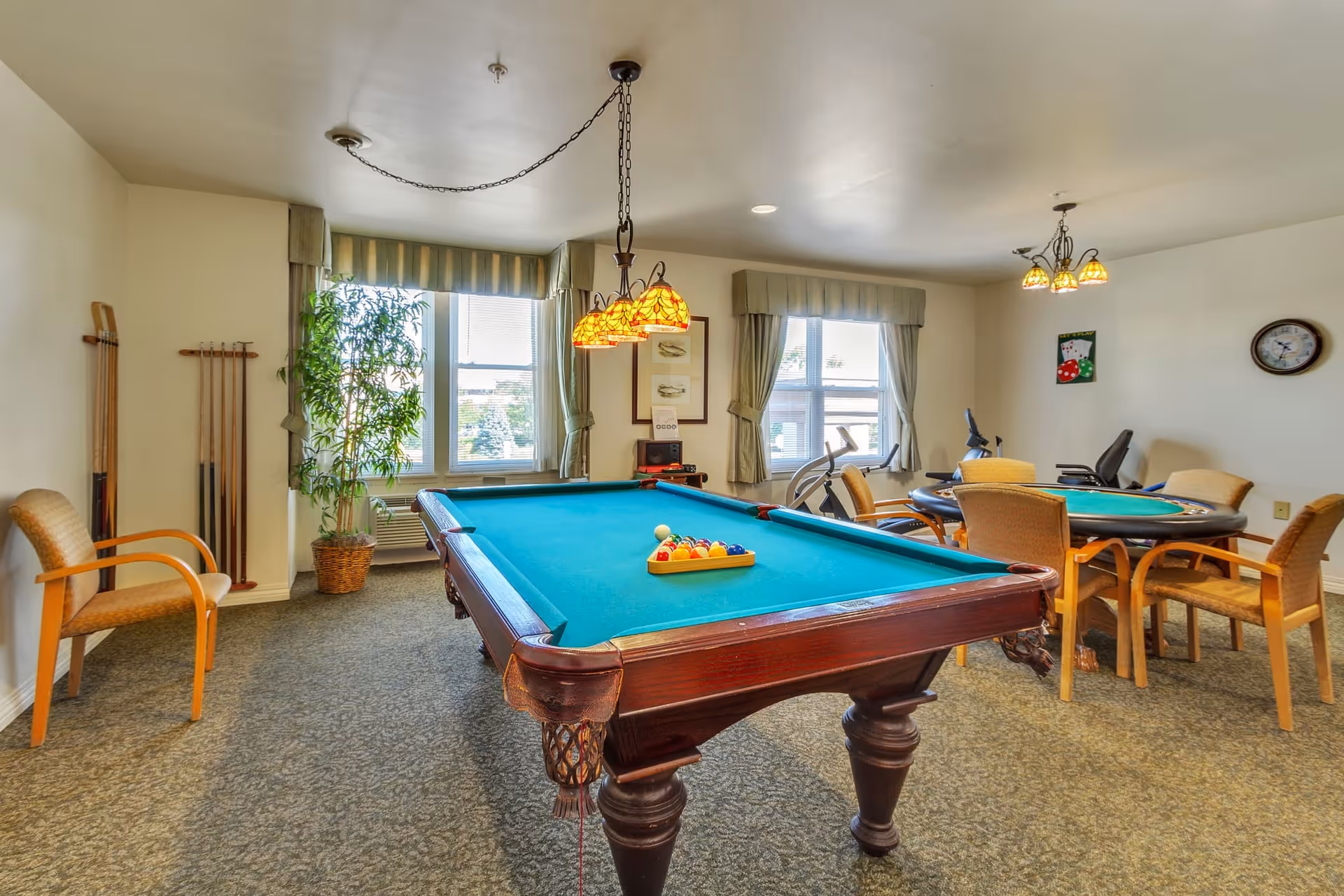 A recreational room with a pool table in the center, a set of pool cues on the wall, a potted plant near the window, a round table with chairs, and exercise equipment near the windows. The room has carpeted floors, two windows with curtains, and ceiling lights with decorative shades.