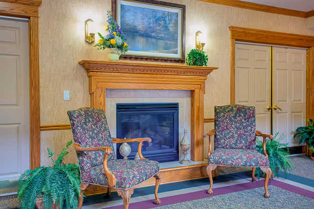 A cozy interior seating area with two patterned armchairs placed in front of a wooden fireplace mantel. The mantel is decorated with a vase of flowers, a green plant, and a framed landscape painting above it. There are wall sconces on either side of the painting, and potted ferns are positioned near the chairs. The room features beige walls with wooden trim and two closed doors in the background.