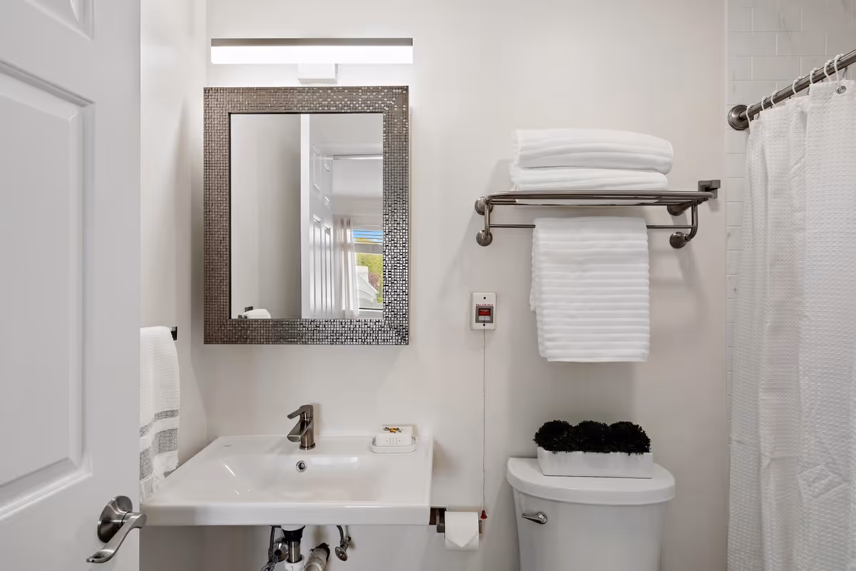 A clean and modern bathroom with a white sink and silver faucet, a rectangular mirror with a mosaic frame above the sink, a towel rack holding folded white towels above a toilet, a small plant on the toilet tank, and a white shower curtain to the right.