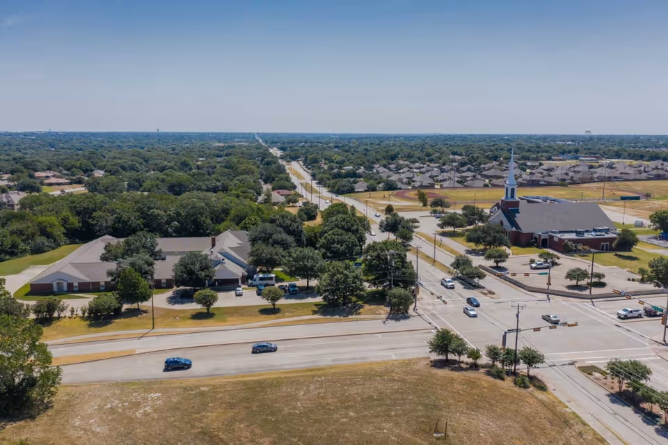 Aerial view of a suburban area showing a senior living facility surrounded by trees and roads, with a church and residential houses in the background under a clear blue sky.